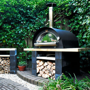 A black sided pizza oven on top of a wooden bench with logs underneath.
