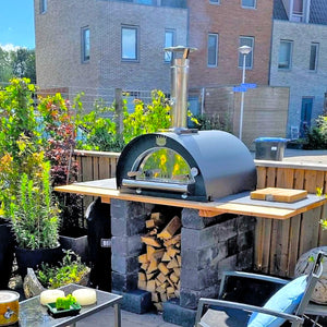 A black sided pizza oven in a low fenced garden with a building in the background and a prep board to the left of the oven.