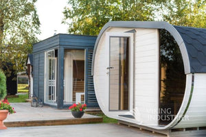 The Delight sauna in white (paint) with black shingle roof on a wooden terrace in a modern garden