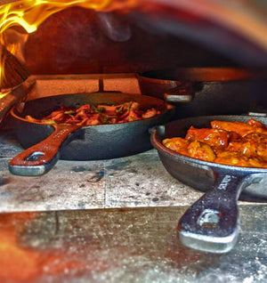 A photograph of the igneus 3 part cast iron set showing the cooking options with an assortment of seasoned chicken dishes, vegetables and marinades these are inside an Igneus oven which is lit currently, logs burning to the left of the oven.