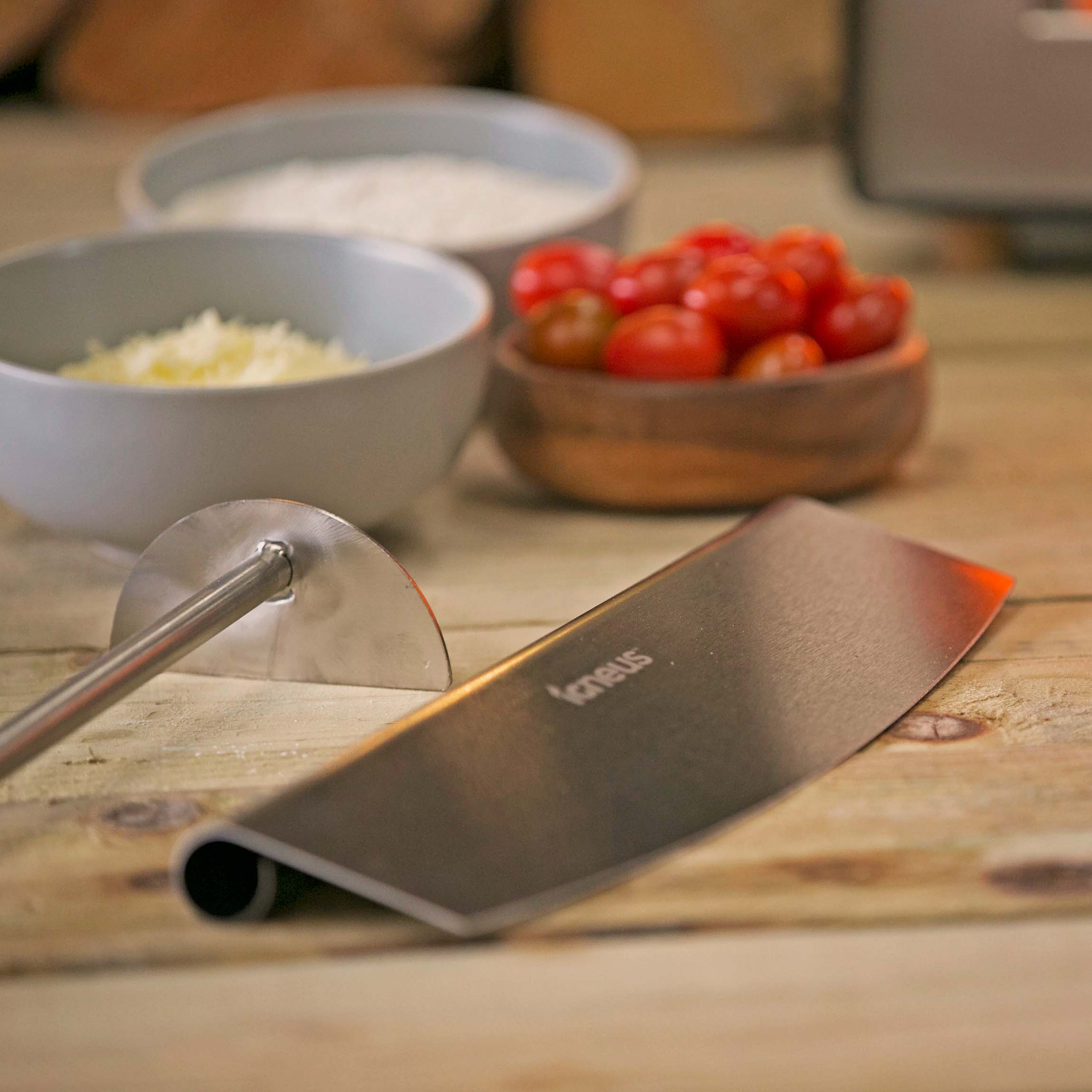 Showing two items from the set in the main focus, the rocker cutter and embers rake both silver and with the Igneus logo, sat on a wooden countertop with some bowls in the background containing cheese, tomatoes and salt.