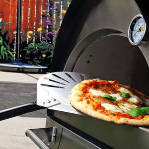 a pizza oven facing left in a garden with flowers in the background - the focus of the image is the Igneus pizza oven peel with a cook pizza atop.