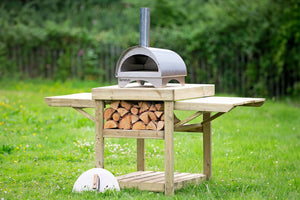 The Minimo outdoor pizza oven atop a wooden stand containing firewood, stood within an open garden