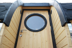A close up image of the sauna barrel door and window with a light oak finish on the wood and black shingles on the roof.