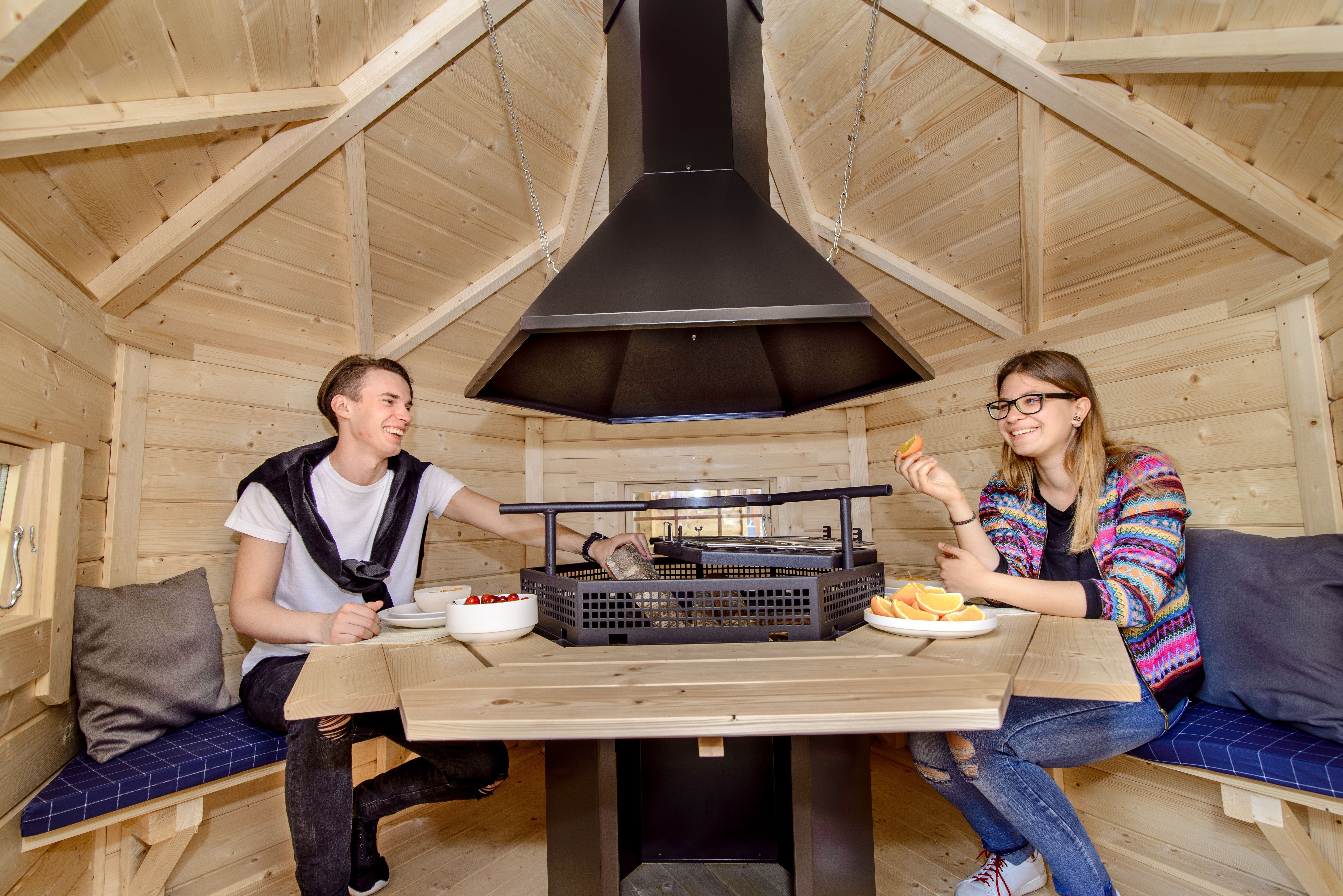 A couple sat inside a grill cabin enjoying some fruit about to light the logs on a Viking BBQ Hut grill.
