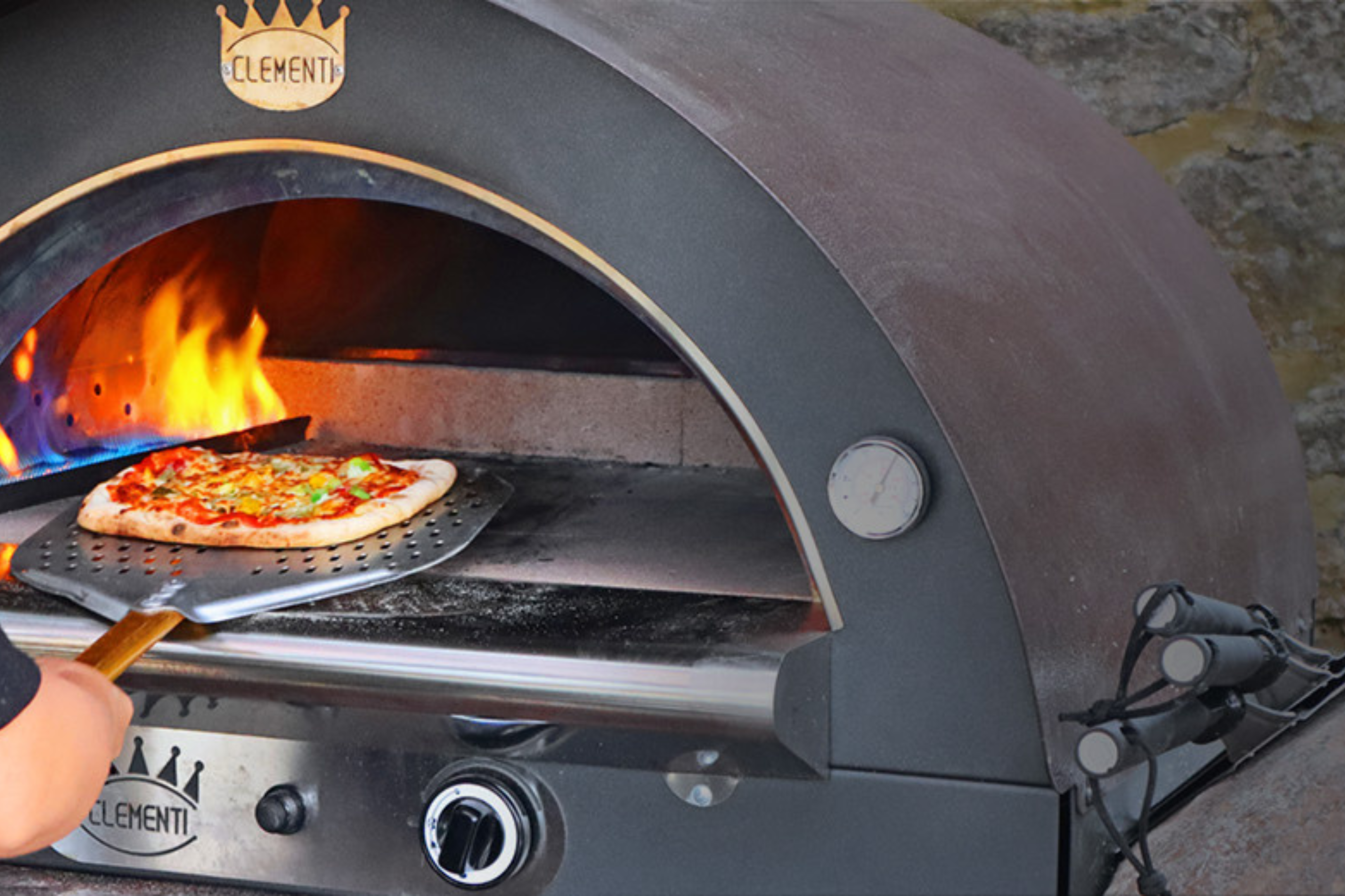 A close up shot of the Gas pizza oven which is lit and a pizza is being pulled out of it with the clementi pizza peel