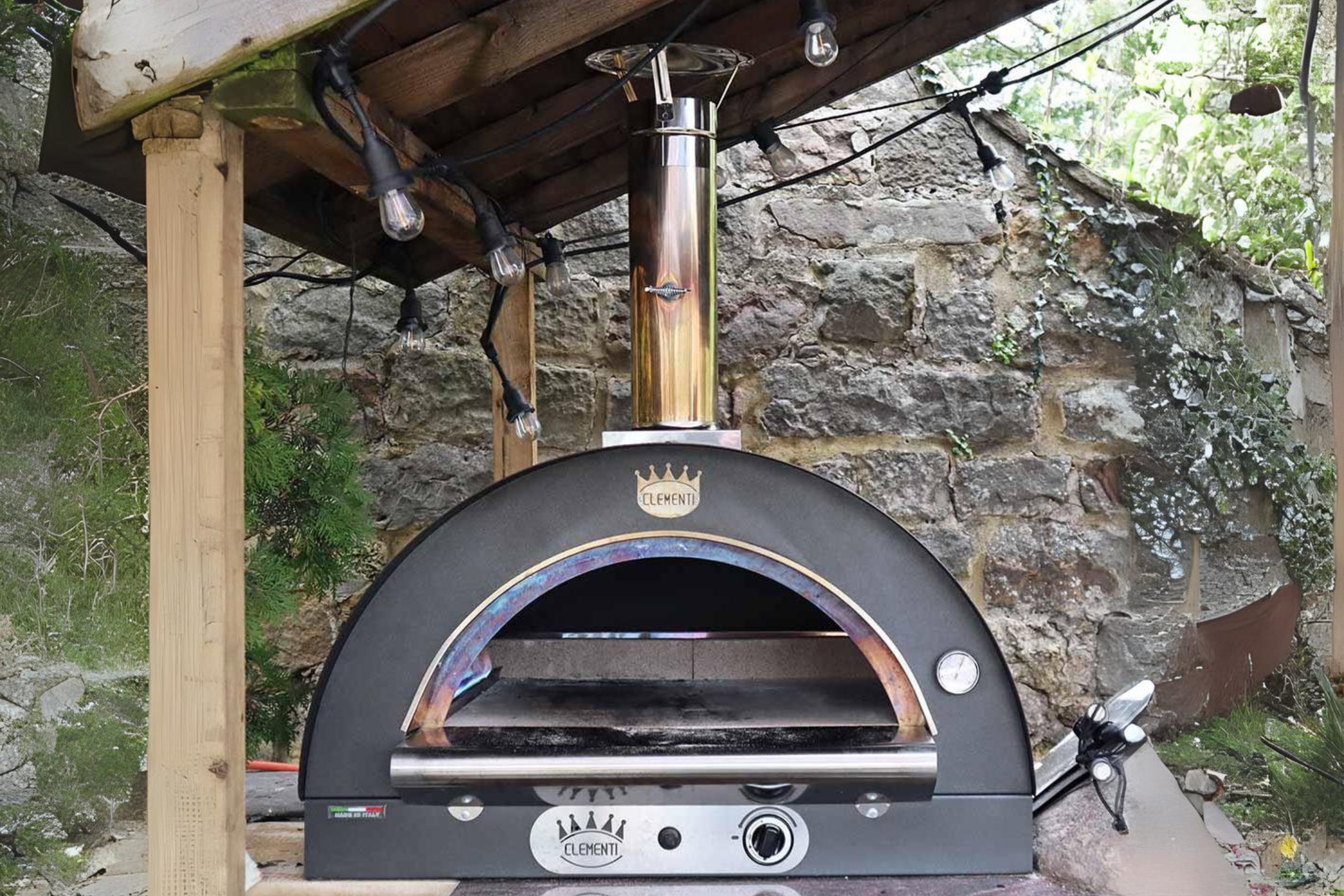 A front shot of a lit Clementi gas pizza oven on a brick surface in a garden undercover.