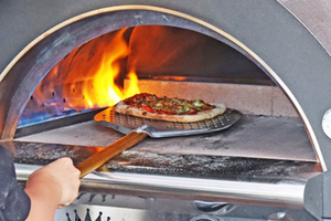 A shot of a cooked pizza being taken out of a lit gas pizza oven outdoor.