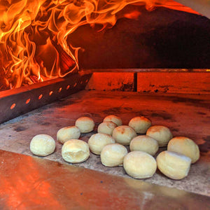 A aclose up shot of some dough balls being cooked inside of a lit woodfired pizza oven.