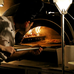 A professional looking chef removing a pizza from a lit woodfired pizza oven.