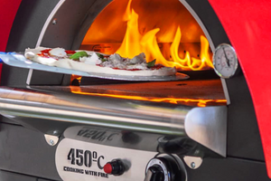 A pizza being placed into a read sided gas fired pizza oven outdoor.