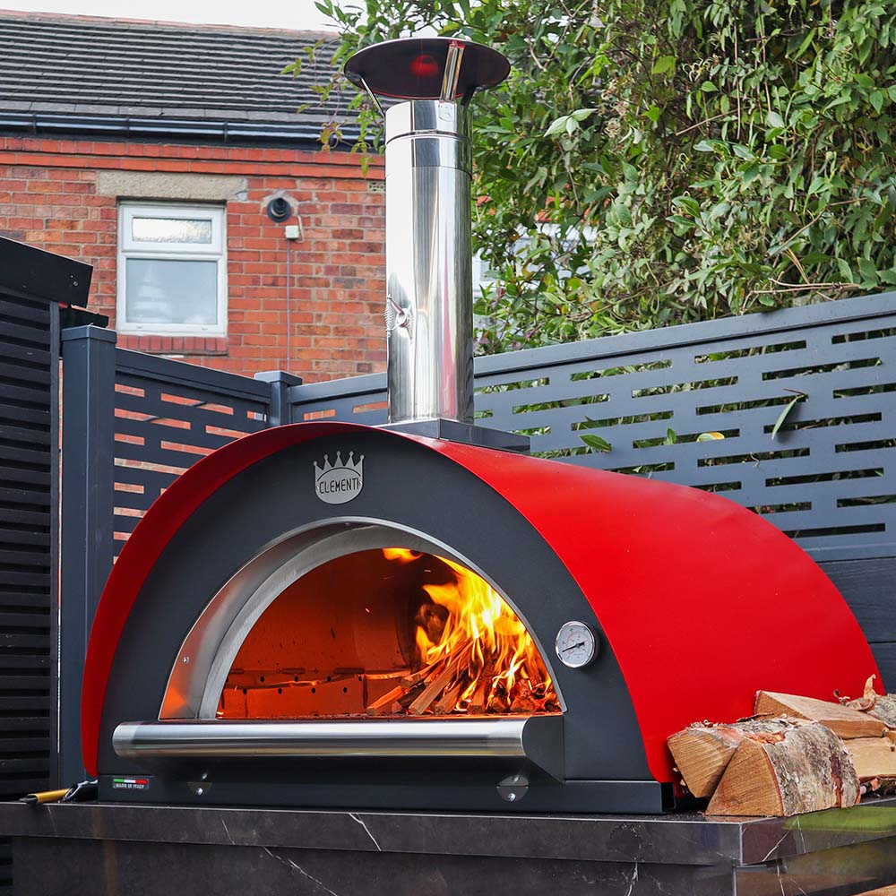 A lit red sided pizza oven with wooden logs to the side of the oven shown in a modern garden with a black fence behind it.