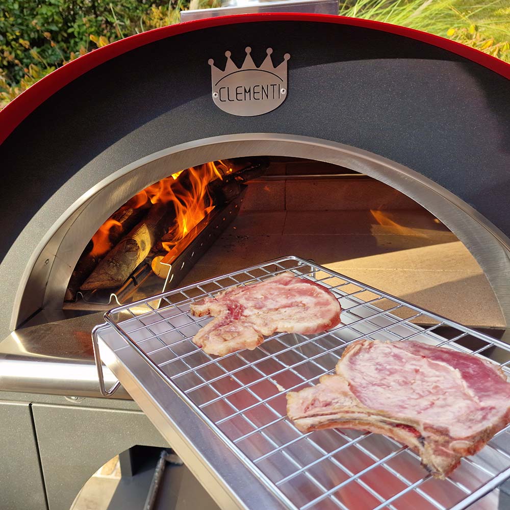 Clementi Pizza Oven, lit with wooden logs burning to the left of the oven, uncooked steaks atop the multicook system about to be placed in the oven