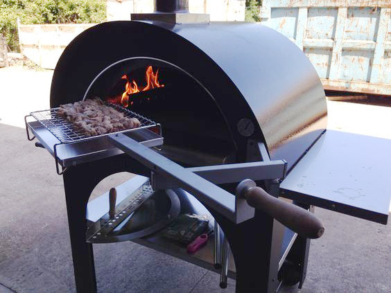 Clementi Pizza Oven, lit with wooden logs burning to the left of the oven, uncooked kebabs atop the multicook system about to be placed in the oven