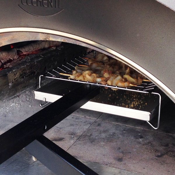 Clementi Pizza Oven, lit with wooden logs burning low to the left of the oven, uncooked kebabs atop the multicook system situated within the oven.