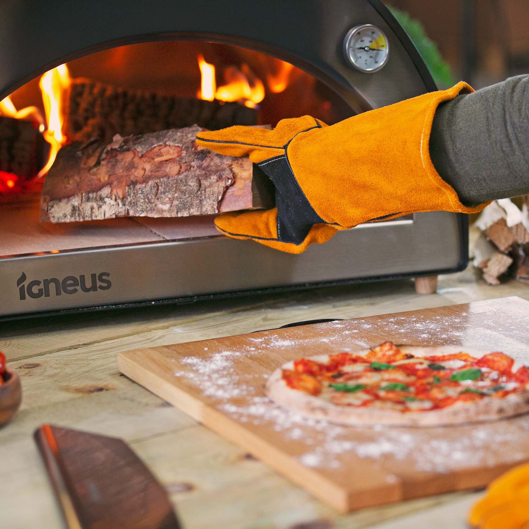 A collection image showing the heat resistant gloves in action placing wood into a lit Igneus pizza oven outdoor. a cooked pizza on a prep board at the front of the image.