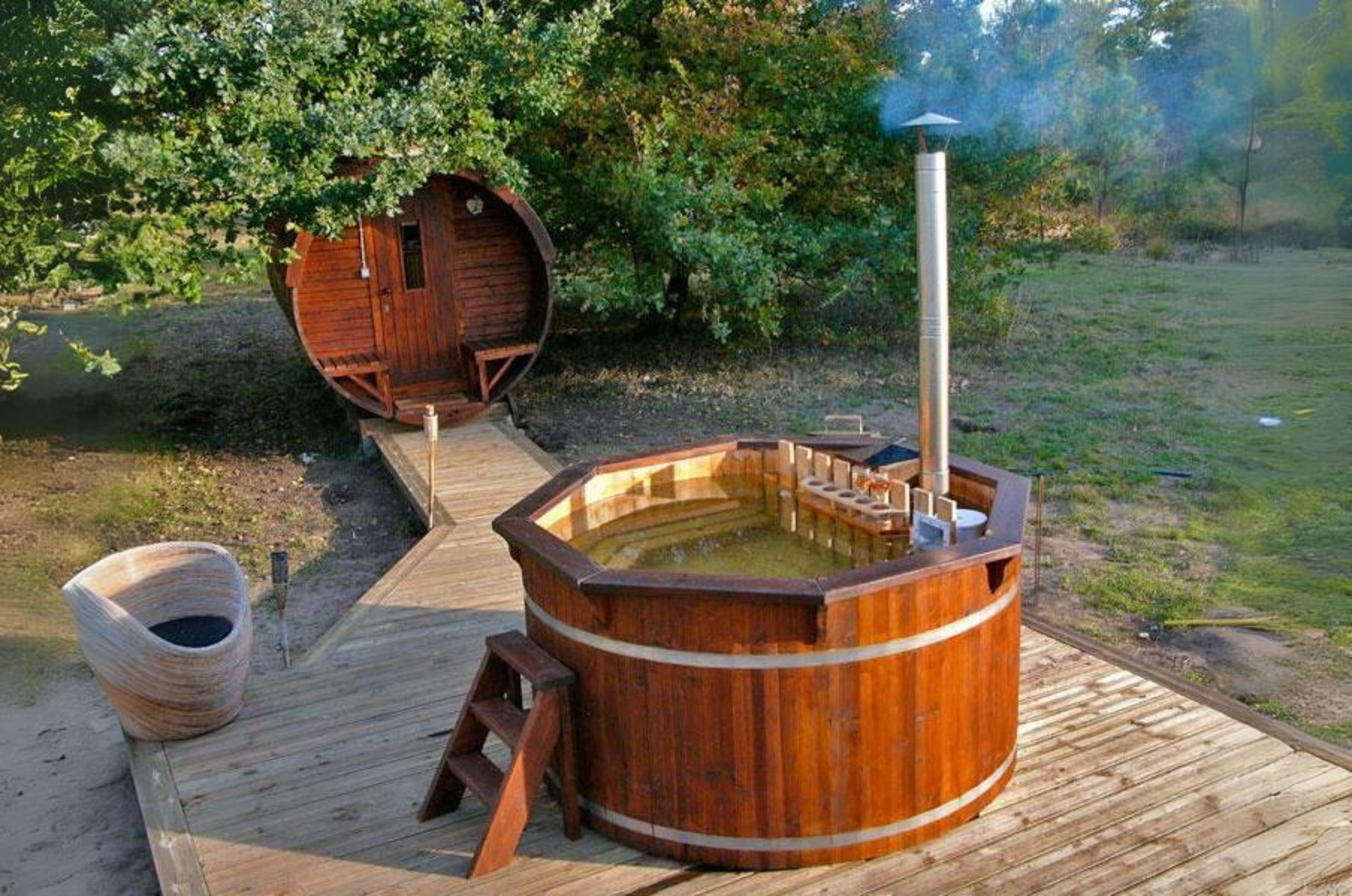 A dark oak treated hot tub with an external heater atop some terrace with a barrel sauna in the background.