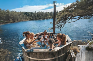 A group of friends sat enjoying the 6 person hot tub sat on a wooden terrace by a lake.