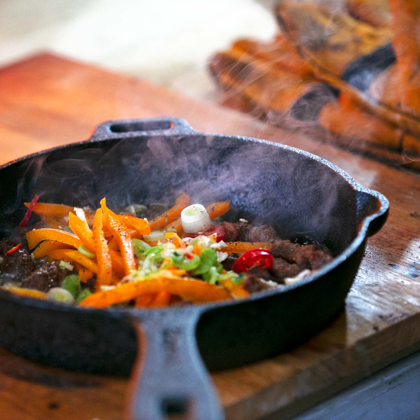 A close up shot of an Igneus cast iron pan with a mixture of meat and vegetables that has been inside of a pizza oven outdoor cooking - currently sat on a chopping board with the orange heat resistant gloves in the background.