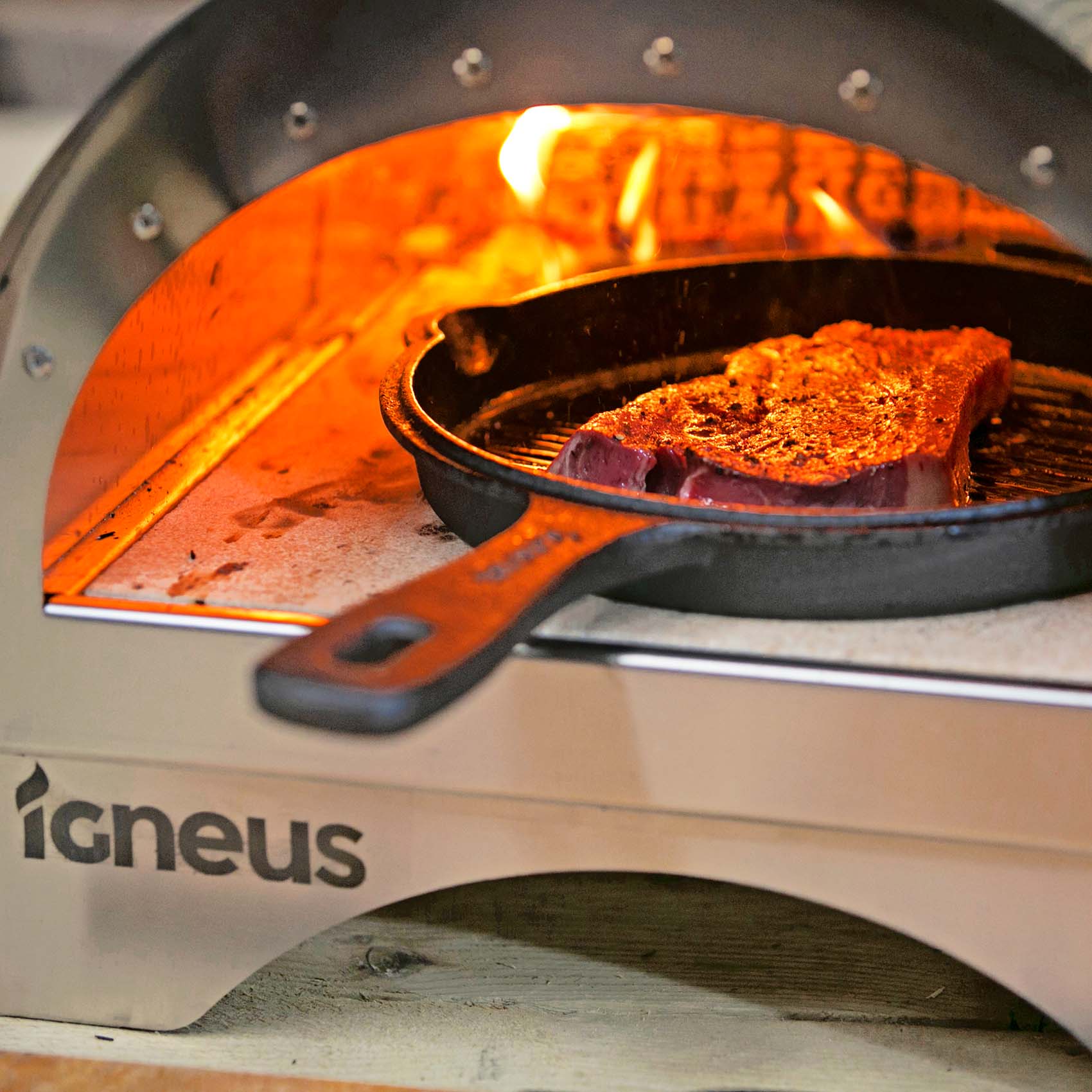 The Igneus Minimo Pizza Oven, silver in colour, lit fire logs at the back of the oven with an black cast iron pan containing a steak cooking away.