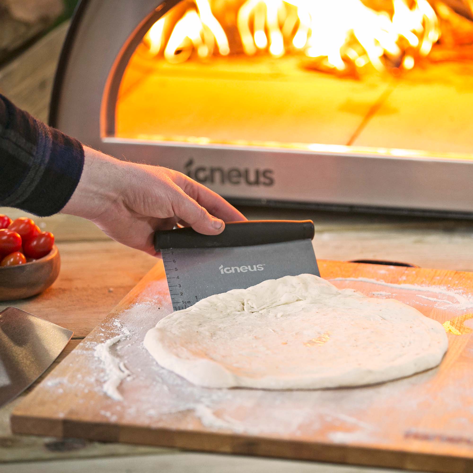 This image shows the Igneus dough cutter in action with some pizza dough on a wooden shopping board in front of a lit Igneus pizza oven. 