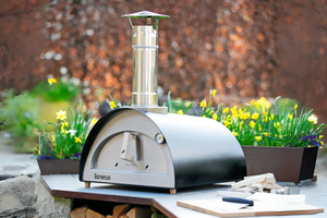 The Bambino bbq pizza oven exterior in matt black sitting atop a wooden bench next to a chopping board in a colourful garden