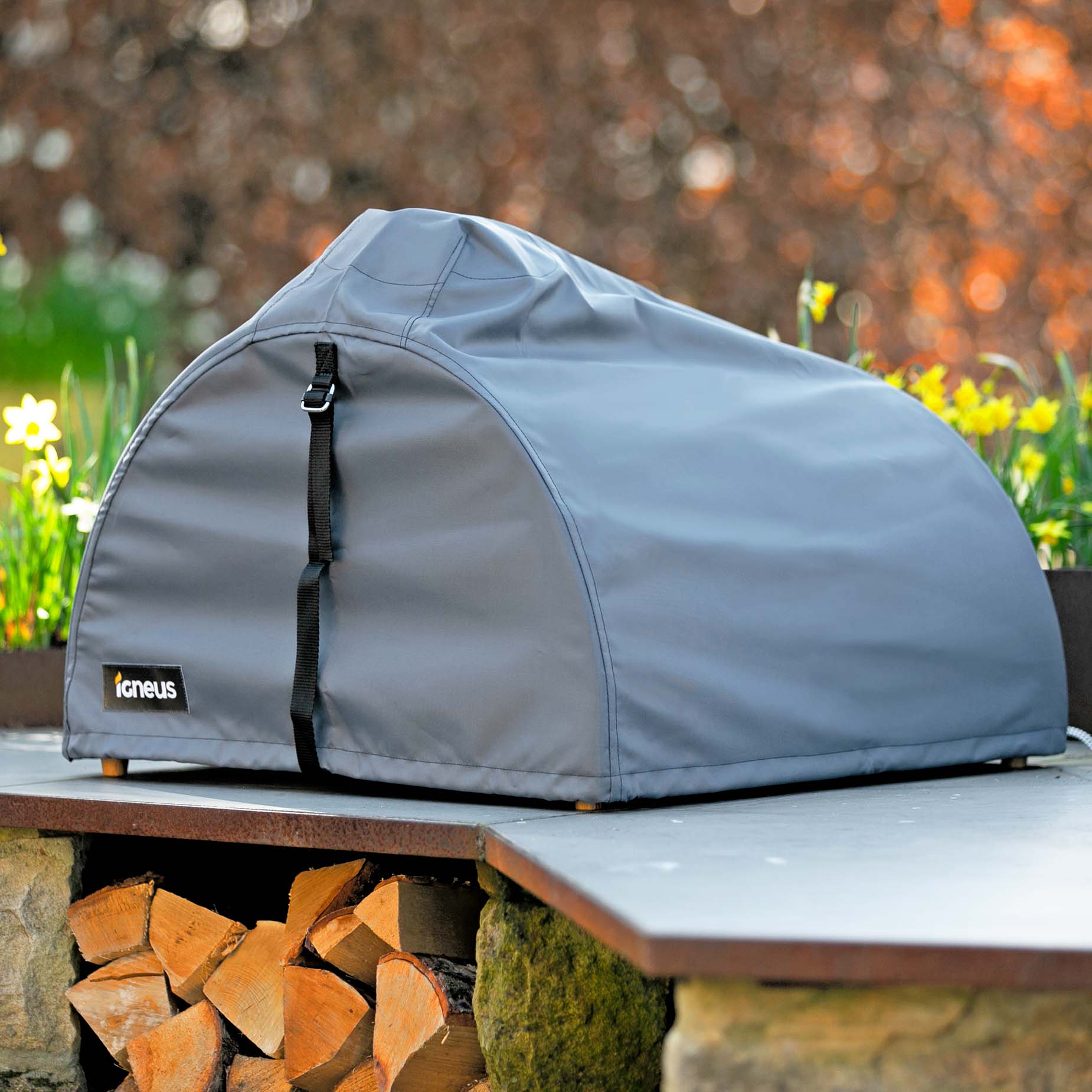 A matte light blue pizza oven cover fixed on top of a BBQ pizza oven on a kitchen counter outdoor with a modern garden in the background and some wooden logs below it.