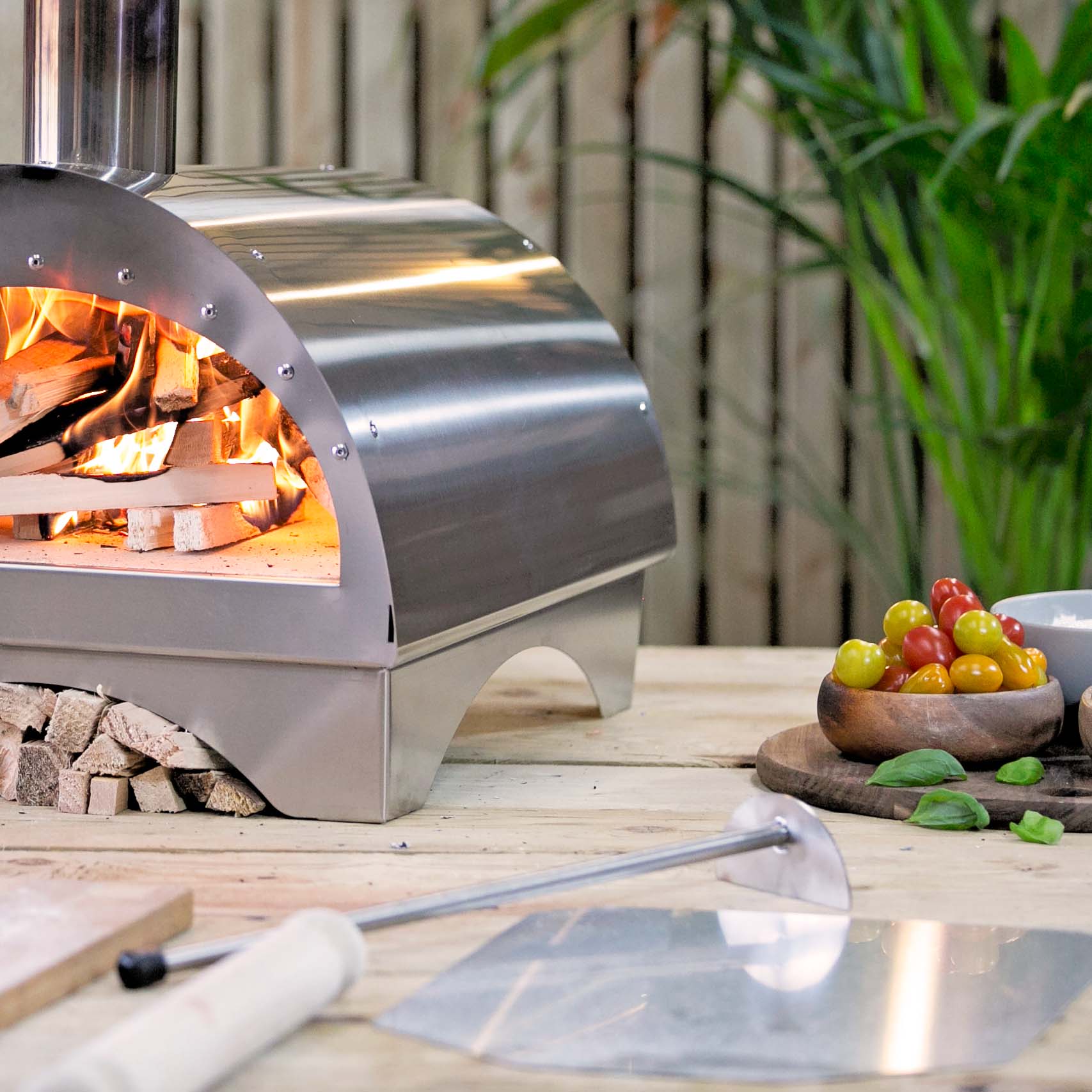 a lit pizza oven to the left of the photo some vegetables and condiments to the right with the Igneus embers rake taking centre stage.