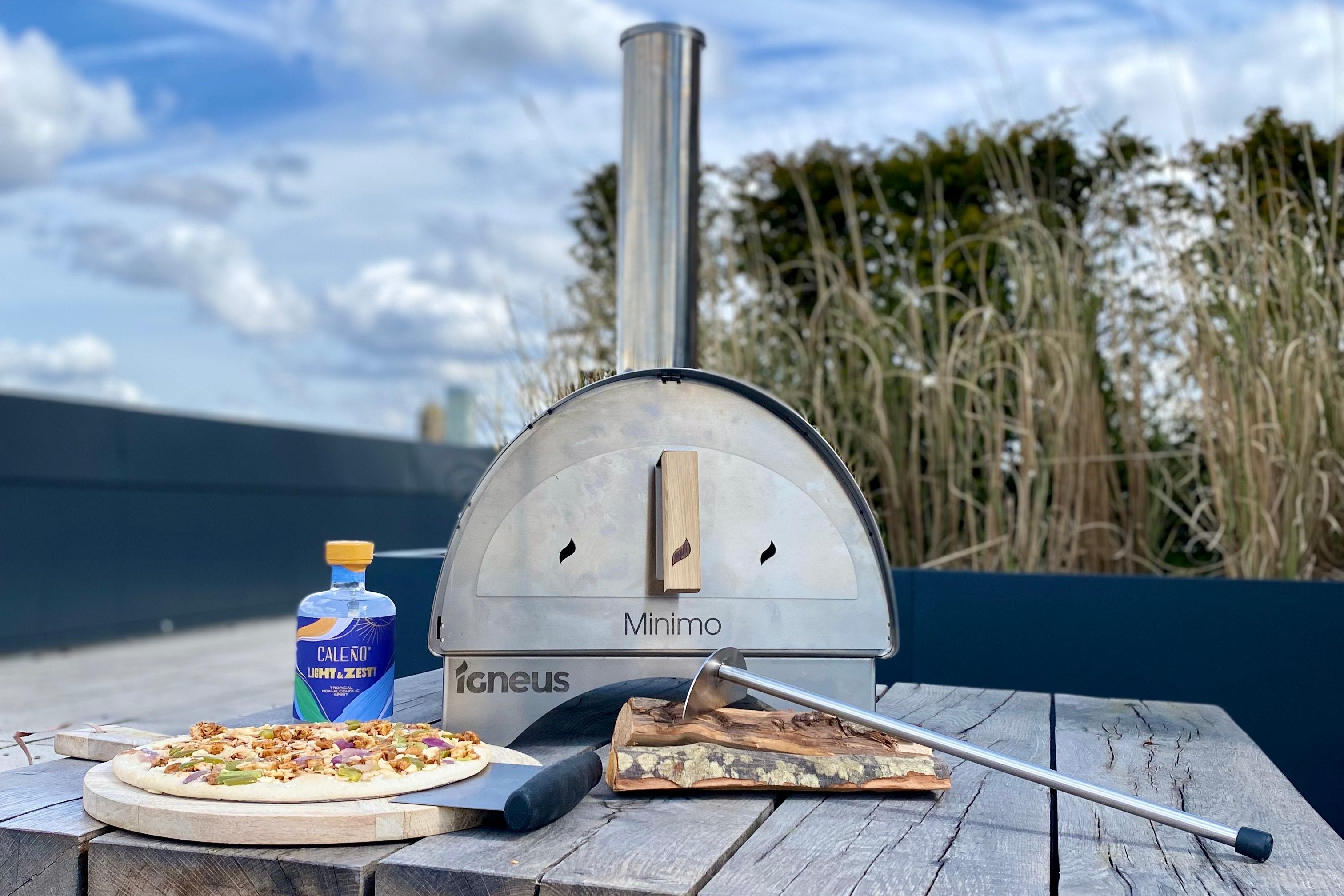 The Minimo wood fired pizza oven outdoor amongst utensils, drinks and pizza atop a wooden table in a coastal garden