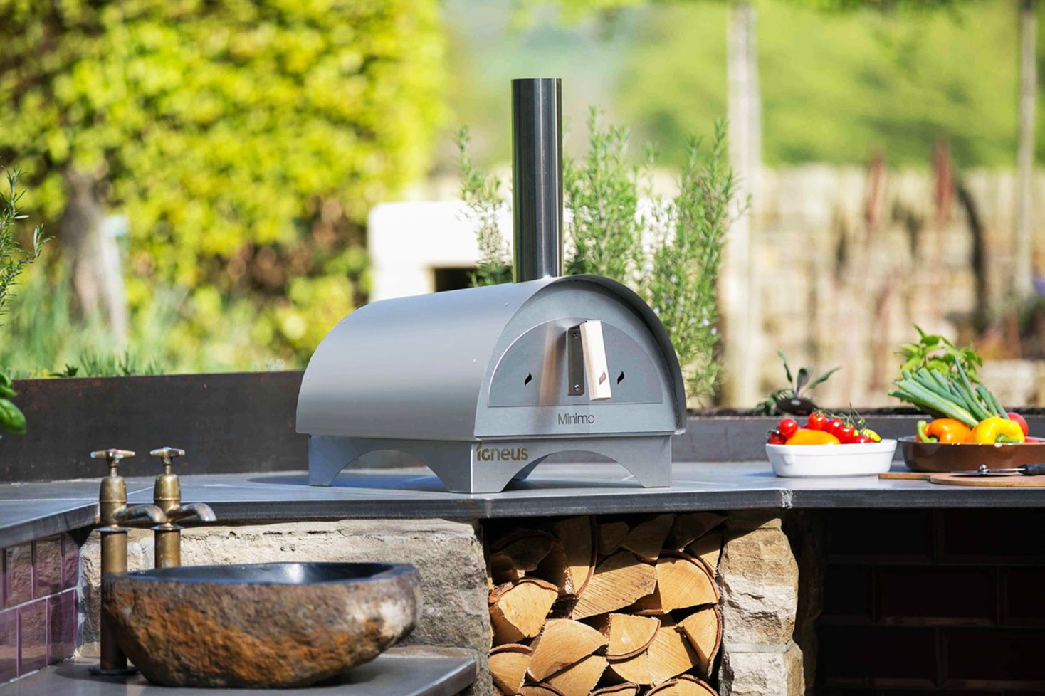 The Minimo bbq pizza oven in a modern garden atop a stone bench with vegetables to the right and a small stone sink to the left