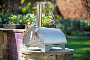 The Minimo wood fired pizza oven outdoor sat atop a stone table in a quaint garden full of flowers