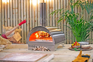 The Minimo garden pizza oven, a homemade pizza cooking within, surrounded by cooking accessories and freshly cut logs