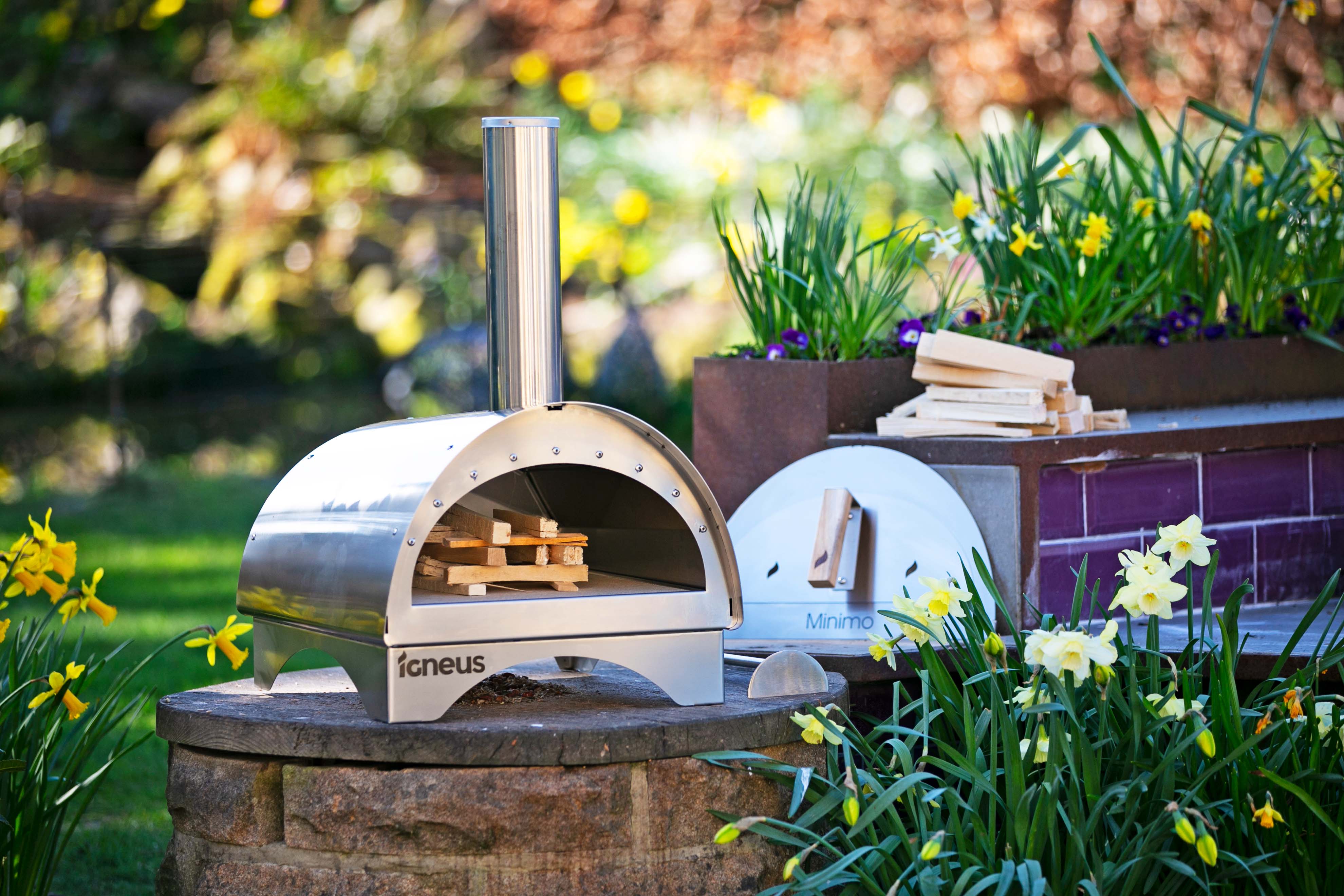 The Minimo bbq pizza oven outdoor sat atop a stone table next to purple brick wall, surrounded by flowers