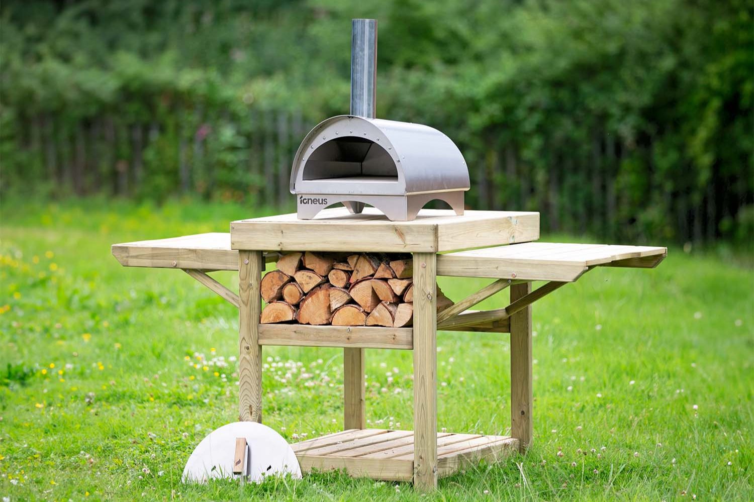 The Minimo outdoor pizza oven atop a wooden stand containing firewood, stood within an open garden