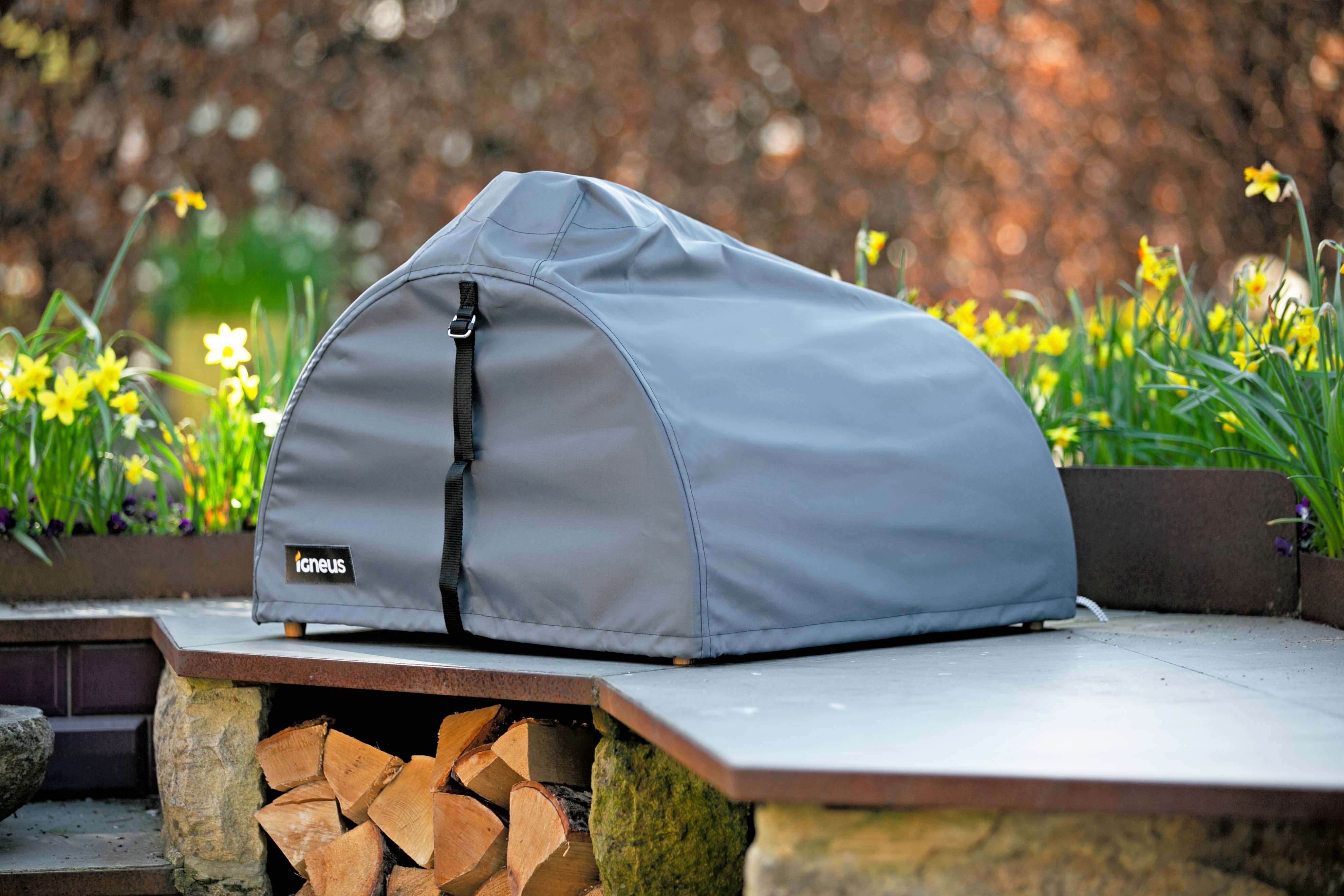 The igneus pizza oven cover in a modern garden logs below and a nice sunny background