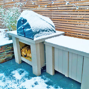 a snowy garden showing the use of the igneus pizza oven outdoor cover with wooden logs below on a countertop and a fence behind.