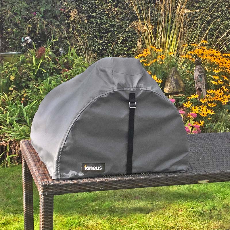 The igneus pizza oven fixed atop a pizza oven on a rattan table in a modern garden with flowers and trees in the background.