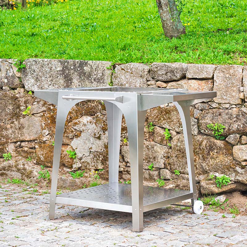 Insitu Silver Pizza Oven Stand Photo - This BBQ pizza oven stand is sat on stone cobbles in front of a brick wall with green grass and a tree in the background.