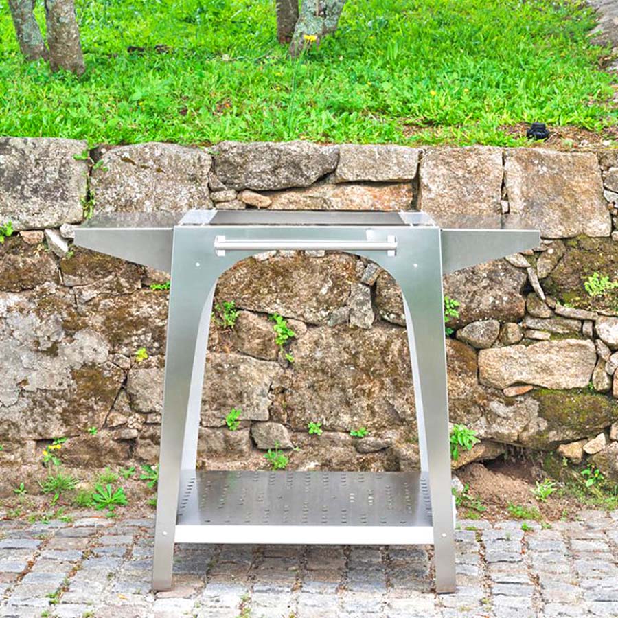 Igneus Pizza Oven Stand - this silver BBQ pizza oven stand in a front on shot is on stone cobbles with grass and trees behind it.