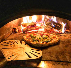 A pizza being spun in a lit pizza oven with the pizza spinner, wooden logs burning in the background.