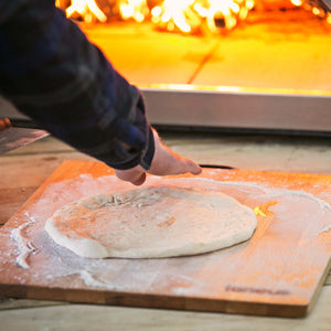 A photo showing pizza dough on top of the igneus pizza oven prep board with a lit pizza oven outdoor in the background.