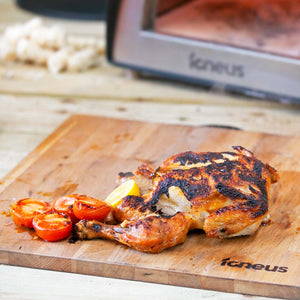 A cooked chicken, roasted tomatoes and a lemon atop the igneus prep board with a BBQ pizza oven in the background.