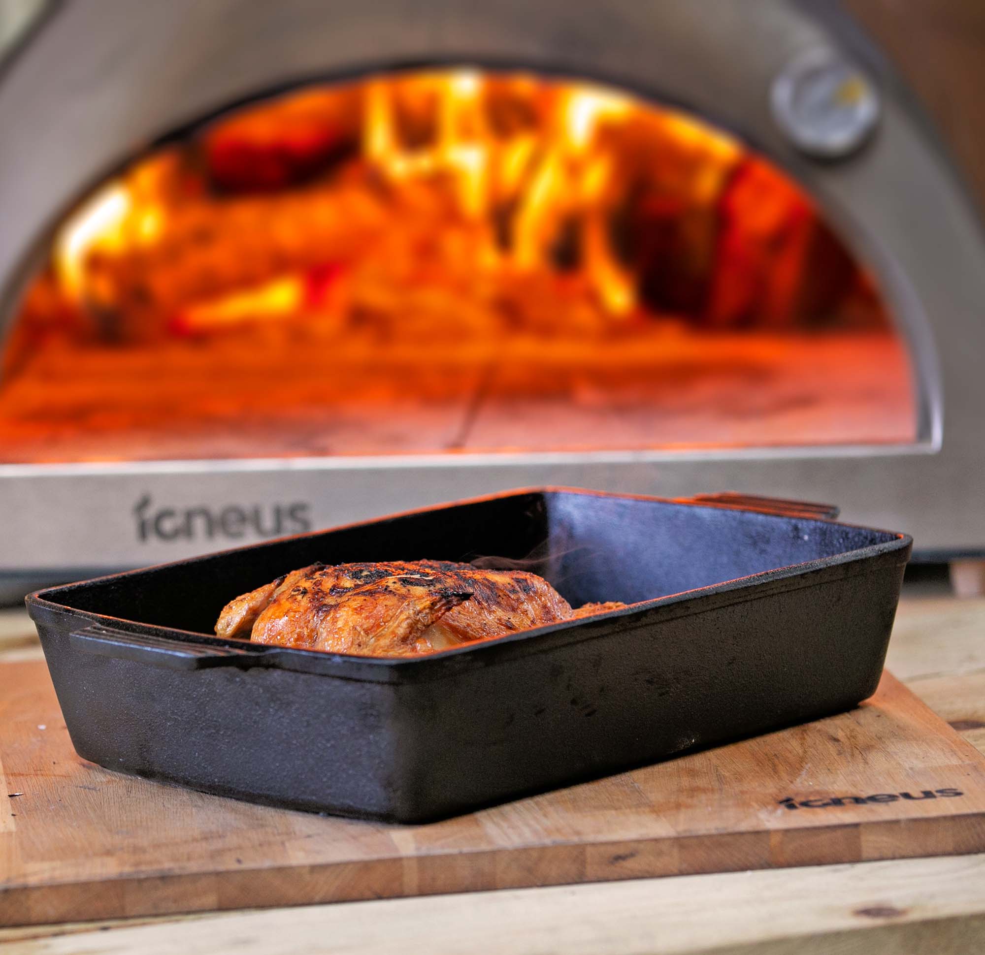 A roasting tray with a cooked chicken atop the Igneus prep board with a lit pizza oven outdoor in the background