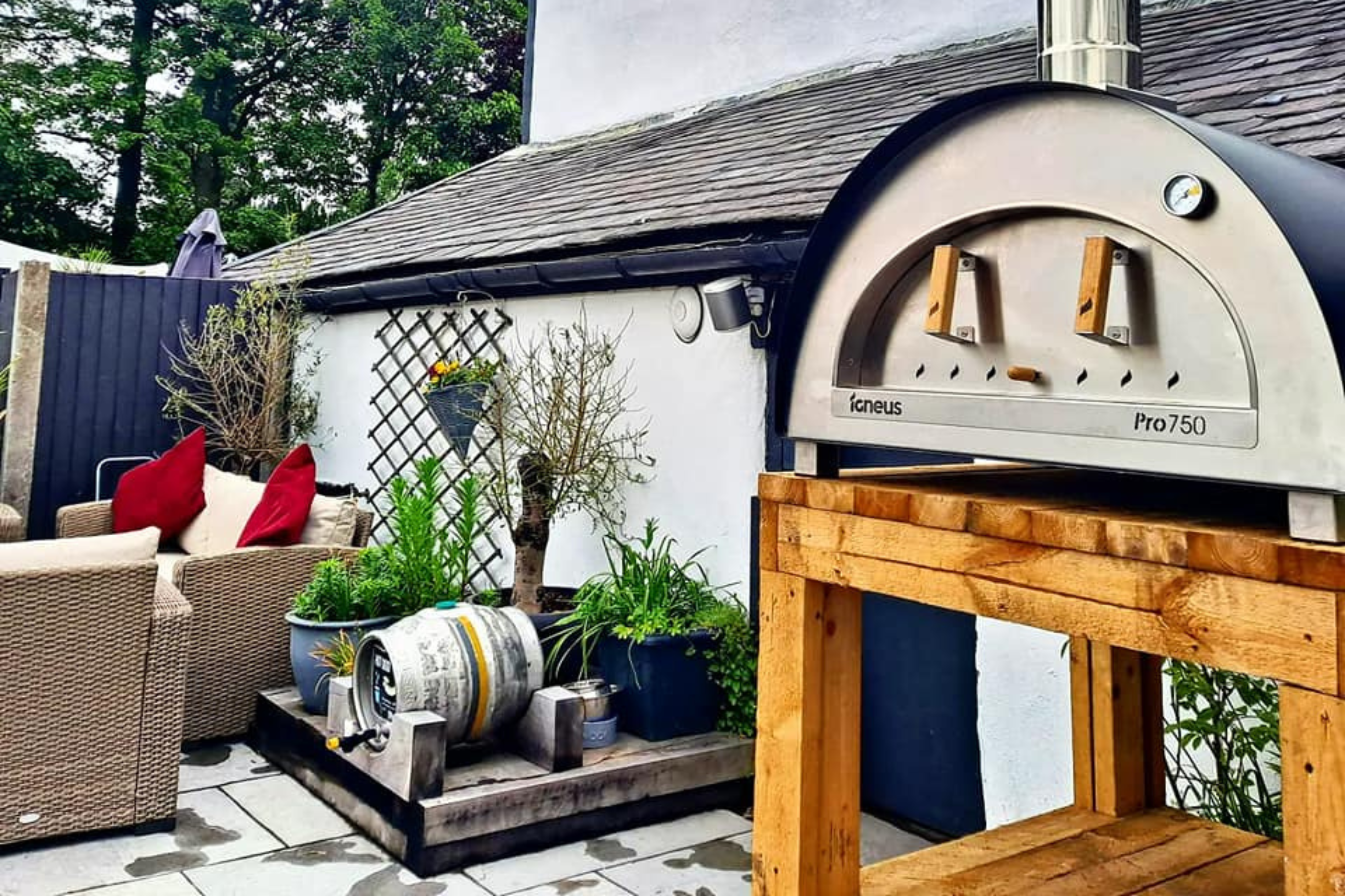 A black sided Igneus Pro 750 Pizza oven outdoor atop of a wooden stand that could be used for wood storage in a pub beer garden with some seating in the background.