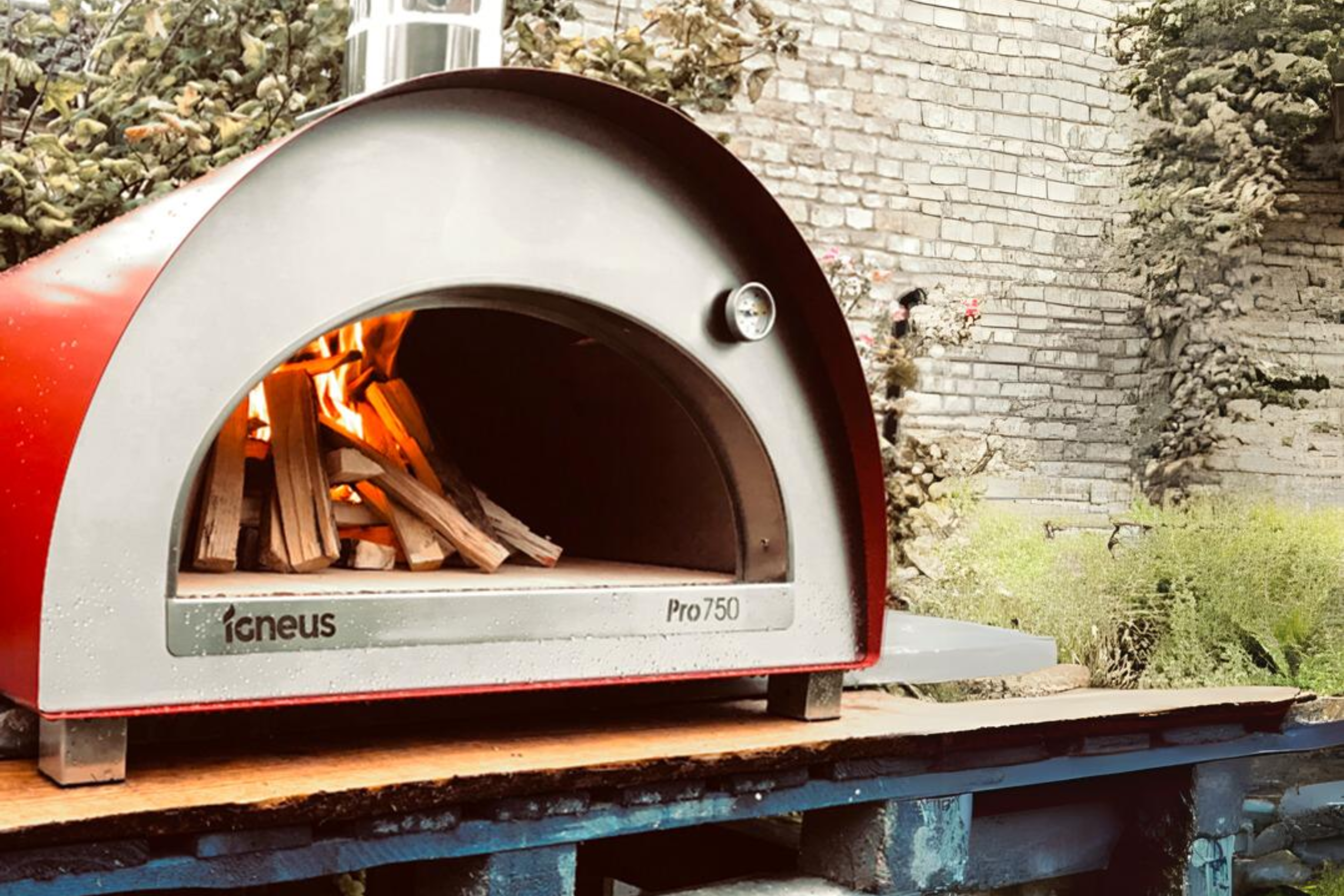 A red sided pizza oven with kindling setting the fire in the oven atop of a wooden surface with a brick wall in the background.