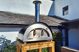 A black sided pizza oven outdoor atop a wooden bench in a garden with a slanted roof behind it, the oven is closed and not lit.