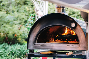 A black sided pizza oven outdoor in a modern garden, the oven is lit with two pizzas cooking inside.