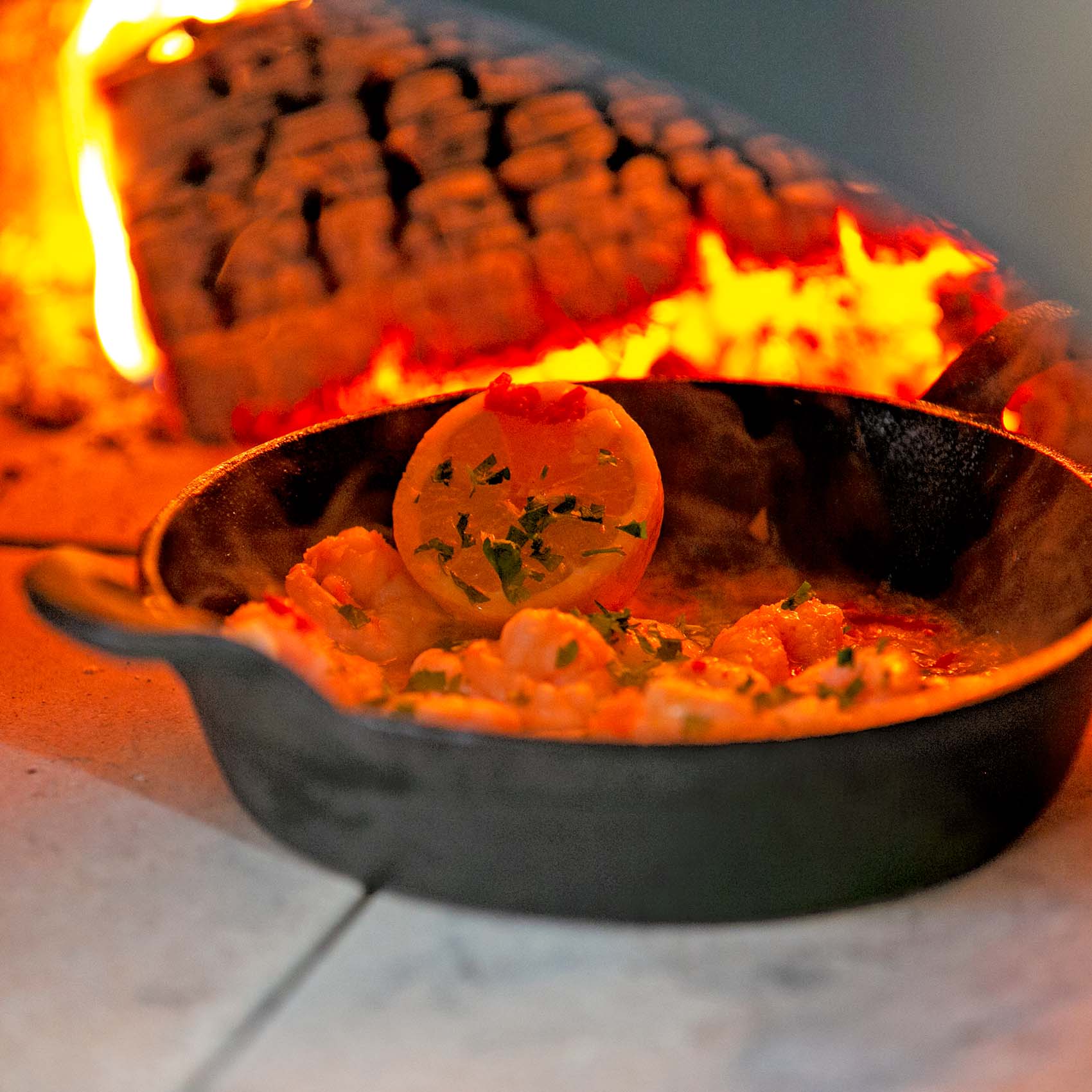 The Igneus pro sizzler dish containing prawns butter and a lemon inside a lit Igneus pizza oven outdoor with a wooden log burning in the background