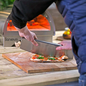 The silver metal rocker cutter is being used to cut a cooked pizza infront of a lit pizza oven