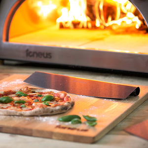 the Igneus rocker cutter is lay next to a cooked pizza in front of a lit BBQ pizza oven